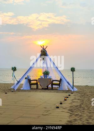 Un cadre romantique pour dîner vous attend sur le sable, avec une verrière magnifiquement drapée et des bougies doucement brillantes. Le soleil se couche sur les eaux calmes de l'océan, créant une ambiance de rêve à Pattaya Thaïlande Banque D'Images