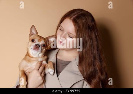 Jolie adolescente aux cheveux rouges avec son chien Chihuahua sur fond beige foncé Banque D'Images