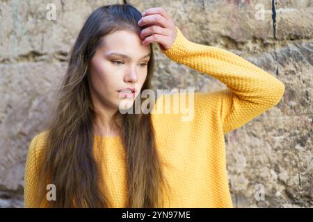 Une femme contemplative vêtue d'un pull jaune vif pose contre un mur de pierre texturé Banque D'Images