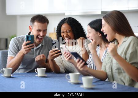 Amis excités célébrant la vérification de leurs téléphones dans la cuisine à la maison Banque D'Images
