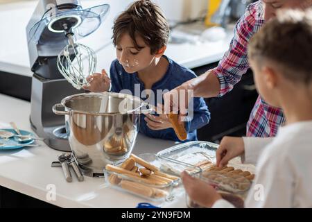 Deux enfants participent joyeusement à une activité de cuisson, mélangeant les ingrédients avec un batteur électrique dans une cuisine lumineuse pendant que l'on goûte la pâte. Banque D'Images
