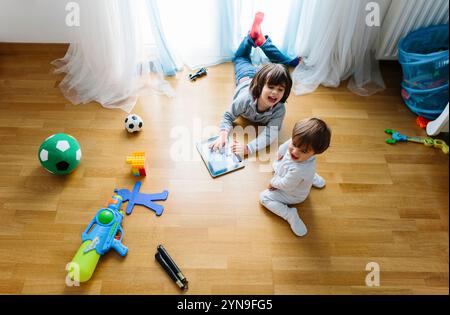 Deux jeunes garçons se livrent à des activités amusantes sur un plancher en bois. Un enfant est allongé sur le ventre avec un livre, tandis que l'autre est assis à proximité, entouré de jouets Banque D'Images