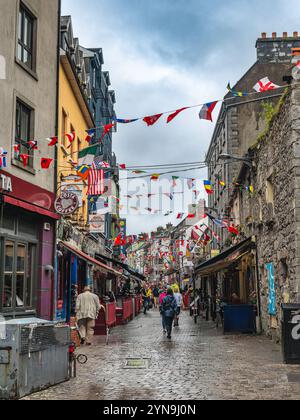 Une rue européenne animée bordée de boutiques et de cafés, décorée de drapeaux colorés. Les gens et les véhicules remplissent la route pavée animée, highlighti Banque D'Images