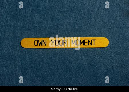 Own Your moment message écrit sur bâton en bois jaune avec fond gris. Symbole conceptuel à l'image de votre moment. Copier l'espace. Banque D'Images
