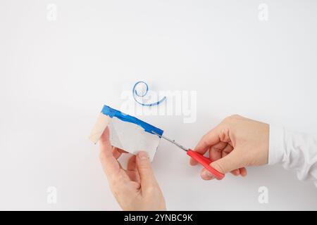 une paire de mains tenant le bord d'un tube en carton avec de la peinture bleue et utilisant une paire de ciseaux à manche rouge pour le couper. Une activité artisanale ou bricolage, partie Banque D'Images