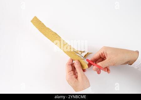 une paire de mains tenant une bande de matériau jaunâtre, carton de boîte à oeufs, et utilisant une paire de ciseaux à manche rouge pour la couper. un motif en zigzag dessiné sur i. Banque D'Images