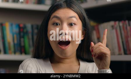 Portrait féminin souriant femme asiatique heureuse étudiante de 20 ans regardant la caméra dans la bibliothèque de l'université montrant les pouces vers le haut comme le symbole d'approbation de geste Banque D'Images