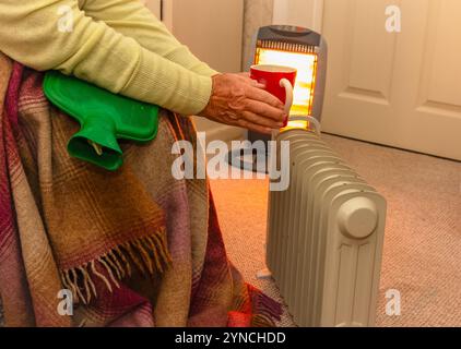 Pensionné essayant de garder au chaud près d'un radiateur rempli d'huile et d'un chauffage halogène avec une bouteille d'eau chaude, une couverture et une boisson chaude. En 2024, le Labour Gover Banque D'Images