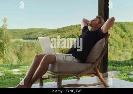 Un homme est assis à l'extérieur avec un ordinateur portable sur ses genoux, profitant d'un moment de détente tout en travaillant à distance dans un cadre naturel. Concept d'équilibre entre vie professionnelle et vie privée Banque D'Images