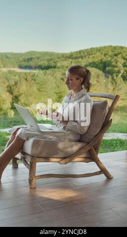 Femme assise à l'extérieur sur la terrasse avec un ordinateur portable, engagée dans une réunion vidéo en ligne ou des conseils, discutant à distance dans un environnement calme et naturel entouré de verdure. Banque D'Images