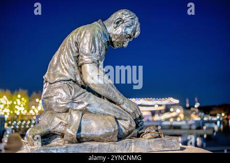 Statue du Maine Lobsterman The Wharf Washington DC | WASHINGTON D.C., États-Unis — Une fonte de bronze du Maine Lobsterman se dresse au quai contre les lumières crépusculaires du front de mer du sud-ouest de DC. Le sculpteur Victor Kahill a modelé l'original de 1939 sur le pêcheur de l'île Bailey H. Bailey Wilbert, montré agenouillé pour accrocher les griffes d'un homard. Un autre moulage de la statue se trouve à Portland, dans le Maine. Banque D'Images
