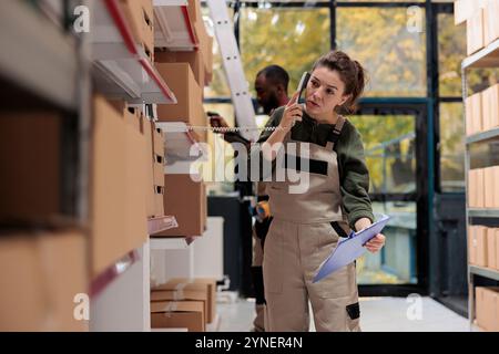 Femme superviseur parlant au téléphone fixe avec le gestionnaire à distance, discutant de l'inventaire des produits dans l'entrepôt. Employé travaillant aux commandes des clients, préparant des boîtes en carton pour l'expédition Banque D'Images