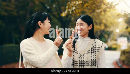 Ville, femmes asiatiques et heureux avec bonbons, voyage et collation pour les amis avec dango, étudiants et la route. Plein air, déplacements et bonbons pour la célébration de Banque D'Images