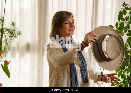 Une femme tenant un chapeau à larges bords, debout dans une pièce légèrement éclairée avec des rideaux transparents. Elle porte une blouse blanche et une écharpe bleue, entourée d'intérieur Banque D'Images