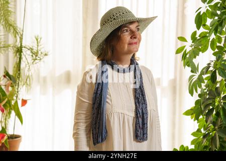 Une femme portant un chapeau de paille à larges bords et une écharpe bleue se tient dans une pièce ensoleillée remplie de plantes. Elle a une expression sereine, avec l naturel doux Banque D'Images