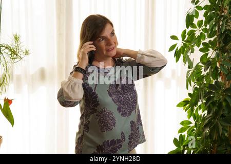 Une femme debout à l'intérieur, souriant tout en parlant au téléphone. Elle a les cheveux bruns aux épaules et porte un haut à motifs floraux. L'arrière-plan f Banque D'Images