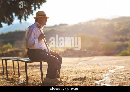 Un homme âgé vêtu d'une chemise blanche et d'un chapeau de paille est assis seul sur un banc, profitant de la lumière chaude du soleil dans un cadre extérieur. Il tient une canne et regarde Banque D'Images