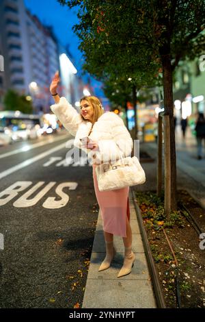 Influenceur à la mode saluant un taxi dans la ville la nuit, tenant son smartphone et un sac à main Banque D'Images