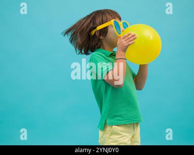 Un jeune garçon dans une chemise verte et des lunettes de soleil jaunes souffle activement un ballon jaune vif. L'arrière-plan présente une teinte bleue fraîche, améliorant Banque D'Images