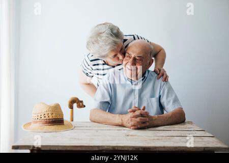 Un couple de personnes âgées partage un moment tendre à la maison, avec la femme embrassant l'homme sur la joue. Ils sont assis à une table en bois, créant un chaleureux et aimant à Banque D'Images