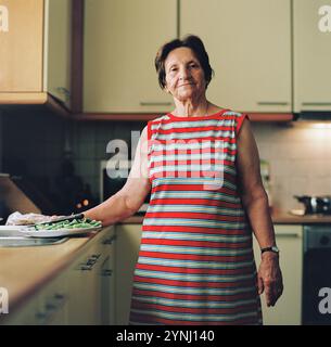 Une femme âgée se tient dans une cuisine bien éclairée, tenant un plateau de légumes frais. Banque D'Images