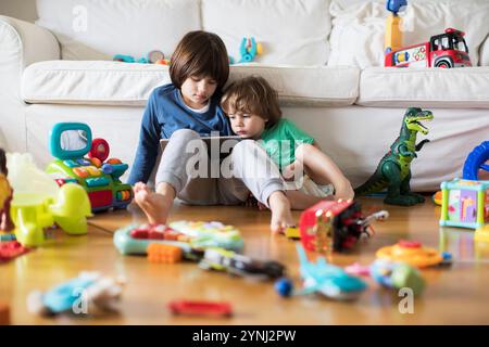 Deux jeunes enfants sont assis sur un plancher en bois dans un salon confortable, engrossé dans un livre. Des jouets colorés sont dispersés autour d'eux, créant un atmos ludique Banque D'Images