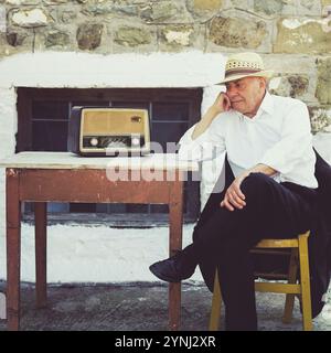 Un homme dans un chapeau de paille s'assoit soigneusement à une table en bois, écoutant une radio vintage placée à côté de lui. L'arrière-plan présente un mur de pierre rustique, Banque D'Images