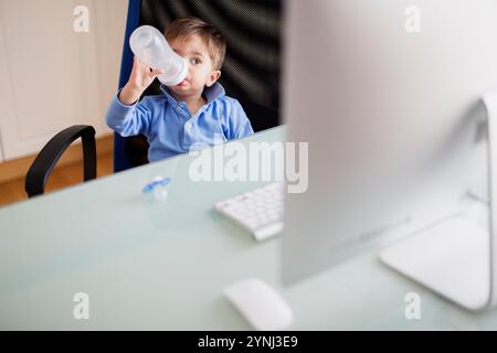 Un jeune enfant est assis dans une chaise à un bureau, sirotant d'une bouteille tout en regardant un écran d'ordinateur. Une lumière naturelle douce remplit la pièce, créant une ambiance chaleureuse Banque D'Images