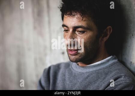 Un homme aux cheveux touffus s'assied tranquillement contre un mur texturé, perdu dans ses pensées. Son expression révèle la contemplation, créant un sentiment d’introspection Banque D'Images