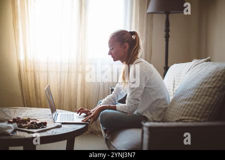 Une femme est assise sur un canapé, concentrée sur son ordinateur portable tout en étant entourée par la lumière douce du soleil filtrant à travers des rideaux transparents. La chambre a une atmosphère chaleureuse et accueillante Banque D'Images