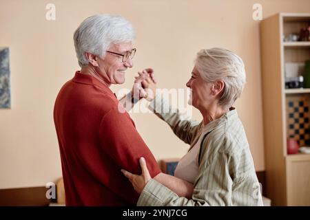 Portrait de vue latérale minimale d'un couple de personnes âgées heureux dansant ensemble et souriant l'un à l'autre en écoutant de la musique à la maison Banque D'Images