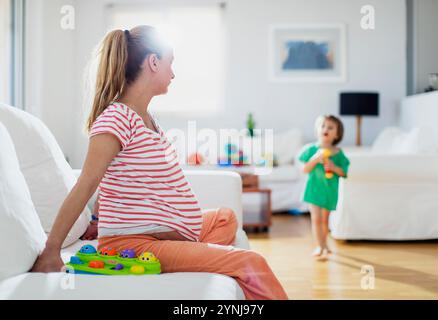 Une femme enceinte s'assoit confortablement sur un canapé dans un salon lumineux, jouant avec un jouet coloré tandis que son tout-petit s'approche avec un expressi joyeux Banque D'Images