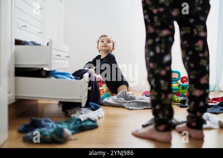 Un jeune enfant est assis sur le sol d'une chambre en désordre, entouré de vêtements et de jouets dispersés. L'enfant lève les yeux, engagé dans le jeu alors qu'il est adulte Banque D'Images