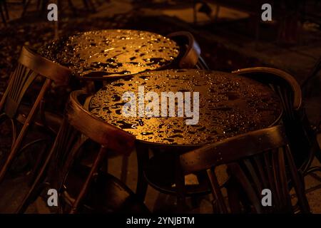 Gros plan de tables de café mouillées et de chaises en bois, illuminées par un éclairage chaleureux et d'ambiance le soir. Les gouttes de pluie brillent sur les plateaux de la table, créant un an serein Banque D'Images