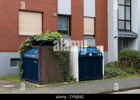 Les bacs de recyclage sont partiellement cachés par des plantes grimpantes à côté d'un bâtiment moderne en briques. Le cadre suggère un environnement urbain respectueux de l’environnement. Banque D'Images