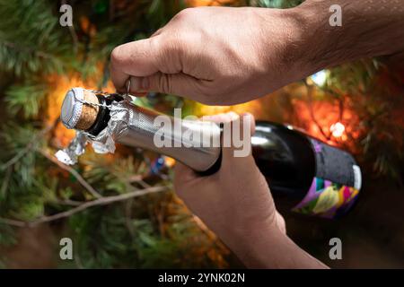 Un jeune homme dans un ouvre une bouteille de champagne sur fond de décorations de Noël, de guirlandes et de lumières Banque D'Images