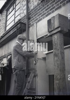 années 1950, historique, à l'extérieur d'un vieux bâtiment, peut-être une vitrine, un ouvrier portant un chapeau plat et avec une cigarette dans la bouche debout sur une échelle en utilisant un morceau de bois pour vérifier le coin de la brique Banque D'Images