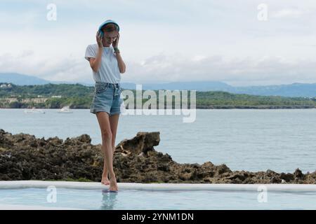 Debout au bord de la piscine, une femme détendue tout en écoutant de la musique sur des écouteurs, profitant de la vue côtière tranquille par une journée douce. Copier l'espace. Banque D'Images