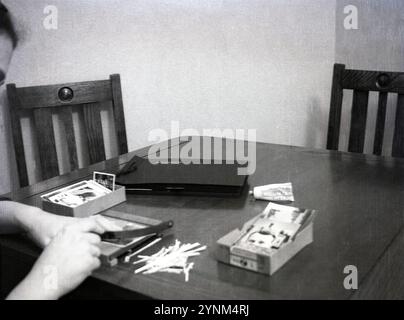 Années 1950, historique, photographie à domicile, assis à une table en bois, un homme utilisant un coupe-papier ou une mini guillotine, pour couper les côtés des photos qu'il mettra dans un album photo, Angleterre, Royaume-Uni. Dans les jours précédant la photographie numérique, les appareils photo utilisaient le film pour capturer des images et pour visualiser les images les tirages devaient être développés dans des laboratoires de films spéciaux à partir des négatifs. Banque D'Images