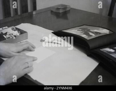 Années 1950, historique, photographie, assis à une table en bois, un homme - mains seulement vues - mettant de la colle sur le dos des photos pour coller à l'intérieur d'un album, Angleterre, Royaume-Uni. Avant l'introduction de la photographie numérique, les appareils photo utilisaient le film pour capturer des images et pour visualiser les images prises, des tirages ou des photos devaient être développés dans des laboratoires spéciaux à partir du film négatif. Banque D'Images