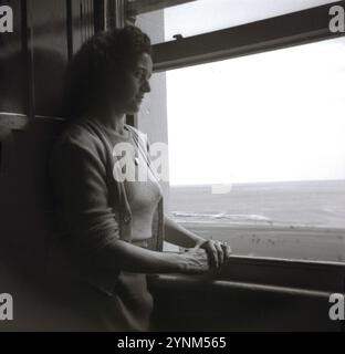 années 1960, historique, une jeune femme, portant un chemisier et un cardigan ouvert, debout près d'une grande fenêtre à guillotine dans la chambre d'hôtel de vacances donnant sur l'océan, la plage et le paysage environnant. Banque D'Images