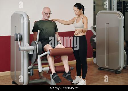 Aider l'homme senior à s'entraîner sur un équipement de gymnastique avec le formateur Banque D'Images