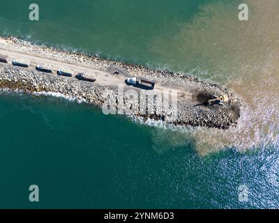 Machinerie lourde et camions vus travailler sur la jetée côtière, entouré d'eau bleue claire dans les heures de lumière du jour. Banque D'Images