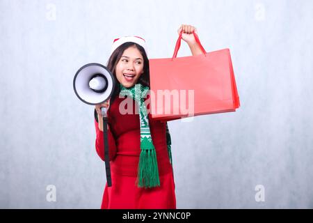 expression de la fille portant un pull de noël rouge avec chapeau de père noël et écharpe, criant joyeusement dans le sac à main tenant haut-parleur mégaphone et portant Banque D'Images