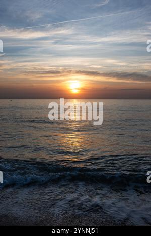 Plan vertical d'un coucher de soleil en mer avec la silhouette d'un phare à l'horizon, de couleur argentée et orange. Banque D'Images