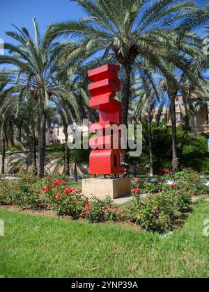 Une sculpture rouge frappante se dresse au milieu de l'herbe verte luxuriante et des roses en fleurs, bordée de palmiers balançant sous un ciel bleu vif. Banque D'Images