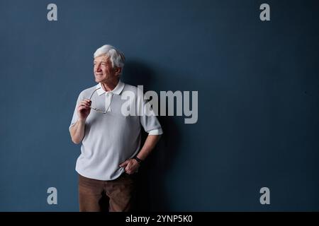 Portrait minimaliste d'un homme âgé aux cheveux blancs portant une tenue décontractée regardant loin debout sur un mur bleu dans un studio avec lumière naturelle et tenant des lunettes Banque D'Images