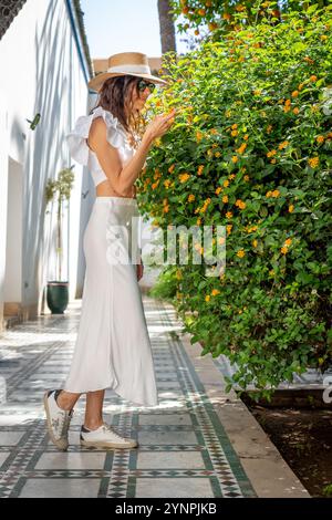 Jeune femme aux cheveux foncés dans chapeau chemise blanche et jupe sentant des fleurs d'orange dans la cour marocaine, Maroc Banque D'Images