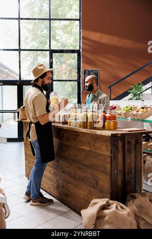 Caucasien commerçant portant un chapeau et un tablier noir tient un pot rempli d'aliments biologiques. Le client regarde autour de lui de l'autre côté du comptoir pendant que le fournisseur dispose les contenants en verre. Banque D'Images