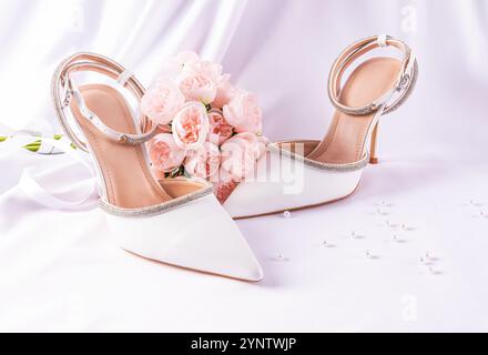 Composition de mariage élégante avec des chaussures blanches chics, bouquet de pivoines roses sur un fond de tissu satiné avec un voile blanc. nature morte. mariée matinale Banque D'Images